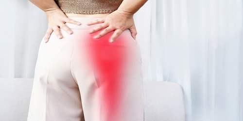 Woman standing facing away, wearing white trousers holding each side of lower waist. A red area in right buttock down back of leg showing sciatic nerve root